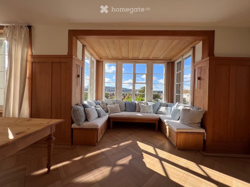 Brightly lit room with wooden floors, large windows, built-in wooden bench seating with multiple cushions, and a wooden table.