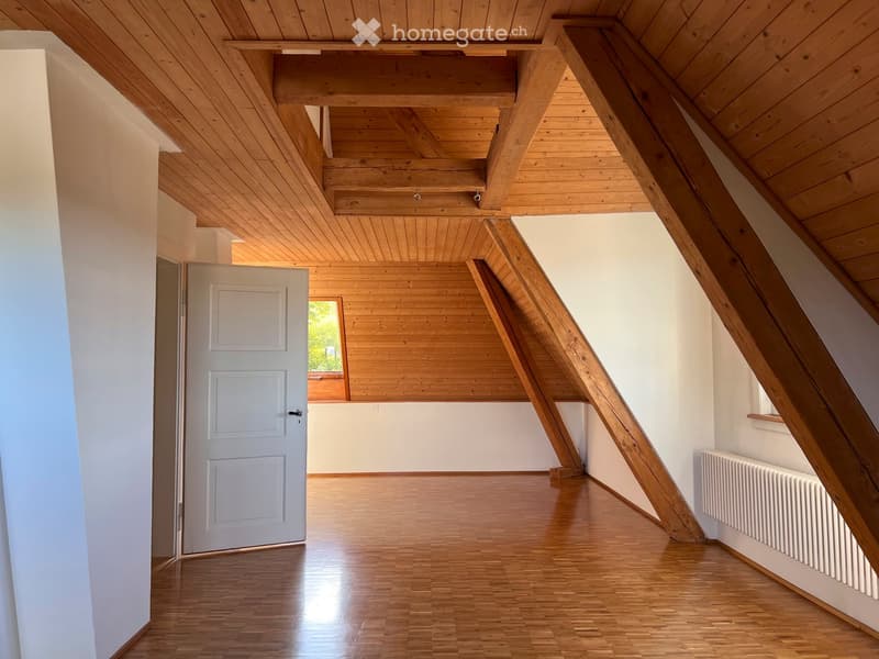 Bright, empty attic space with wooden beams, wooden flooring, white walls, and a white door leading to another room.
