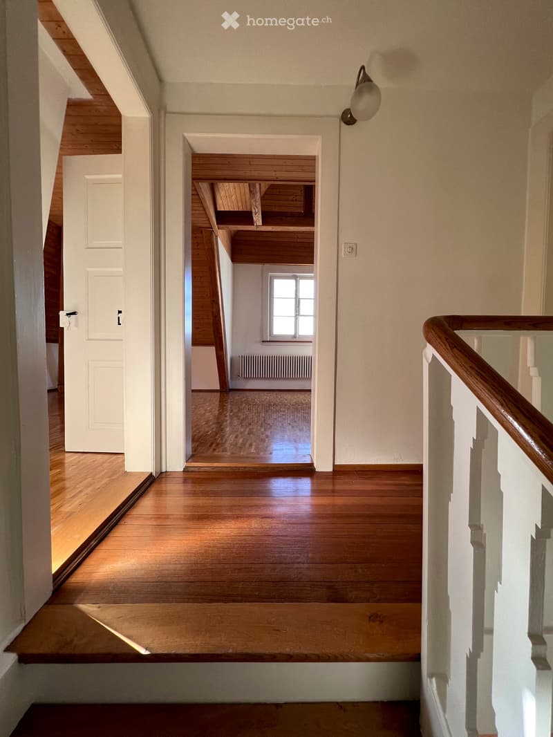 Spacious hallway with wooden flooring, white walls, and a staircase leading upwards. Features a wall-mounted light fixture and a door leading to another room.