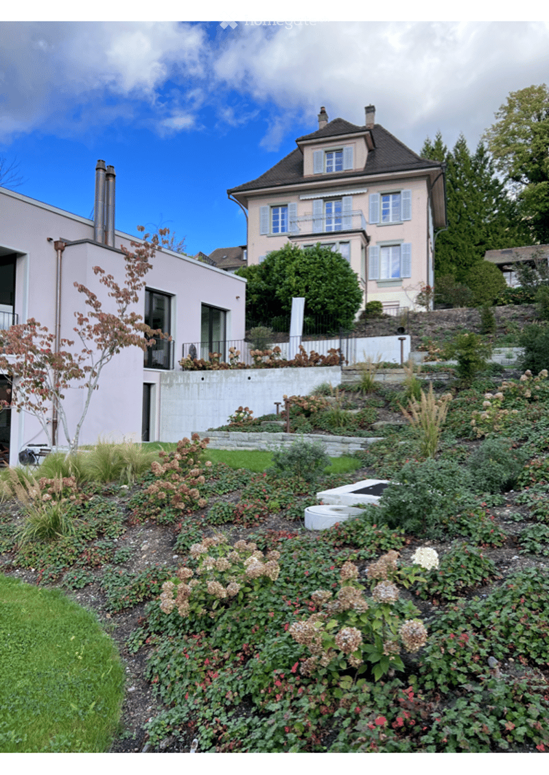 Well-maintained garden with various flowering plants, shrubs, and trees. Includes a modern house with a balcony and a contemporary building with large windows.