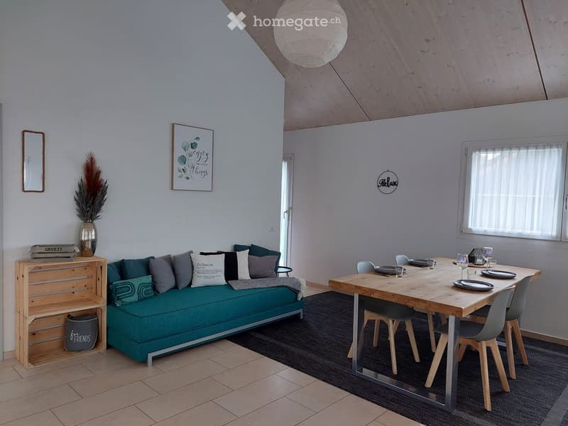 Living room with blue sofa, pillows, dining table with chairs, wooden ceiling, wooden flooring, wooden shelf, vase with plant, table setting, wooden framed window, lamp
