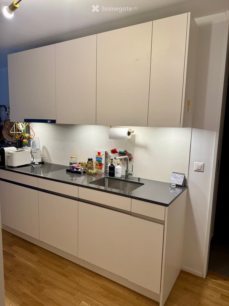 Modern kitchen with white cabinets, black countertop, sink, and various appliances including a toaster and blender. Wooden floor and bright lighting.