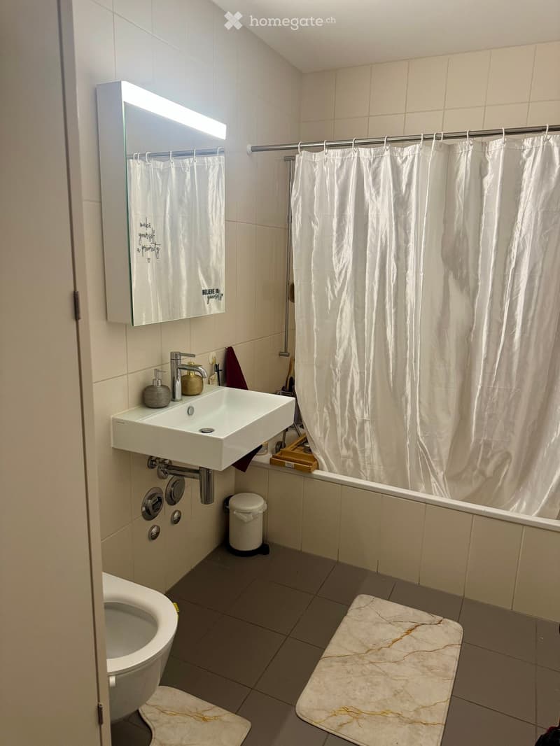Modern bathroom with white tiles, a sink with a modern faucet, a mirror with built-in lighting, a toilet, a bathtub with a white shower curtain, and marble-style rugs on the floor.