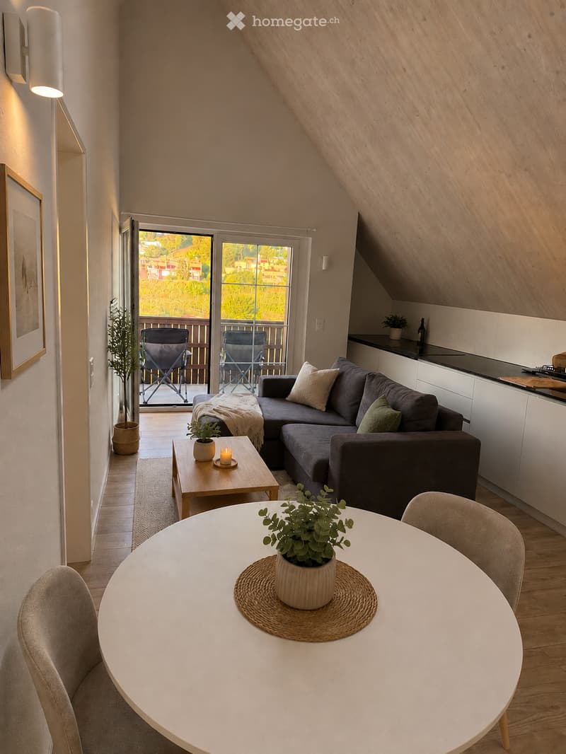 Bright, open living and dining area with wooden floors, a grey sectional sofa, a round dining table with chairs, and a small balcony with outdoor chairs.