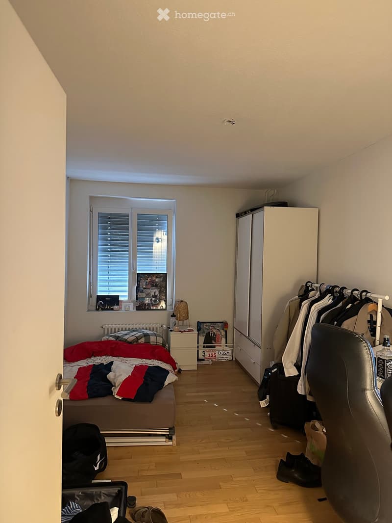 A furnished bedroom with a bed, a chair, a refrigerator, a wardrobe, a nightstand, a laptop, a picture frame, a radiator, a bottle, a handbag, and shoes.