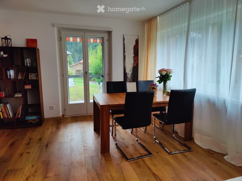 Dining room with wooden floor, large window with curtain, dining table with four chairs, shelf with books and decorative items, vase with flowers on table.