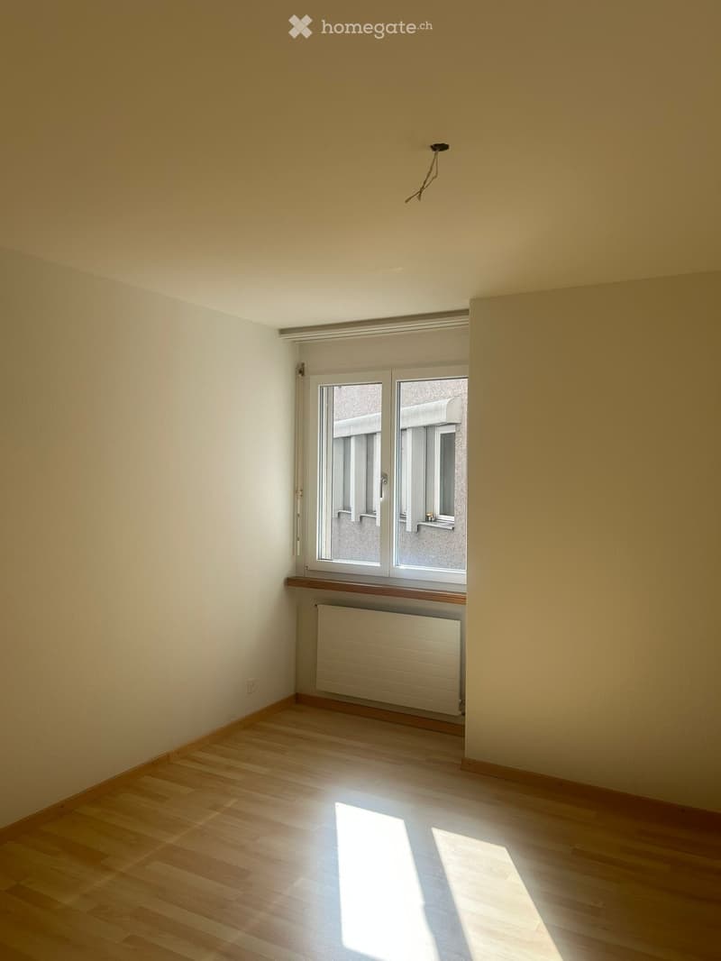 Empty room with light wooden flooring, white walls, and a window with white frames and blinds. A radiator is visible below the window, and the room has a simple, uncluttered design.