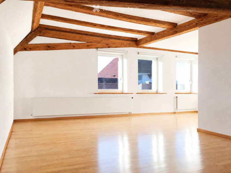 Empty room with wooden floor, white walls, exposed wooden beams on the ceiling, and large windows with white frames.