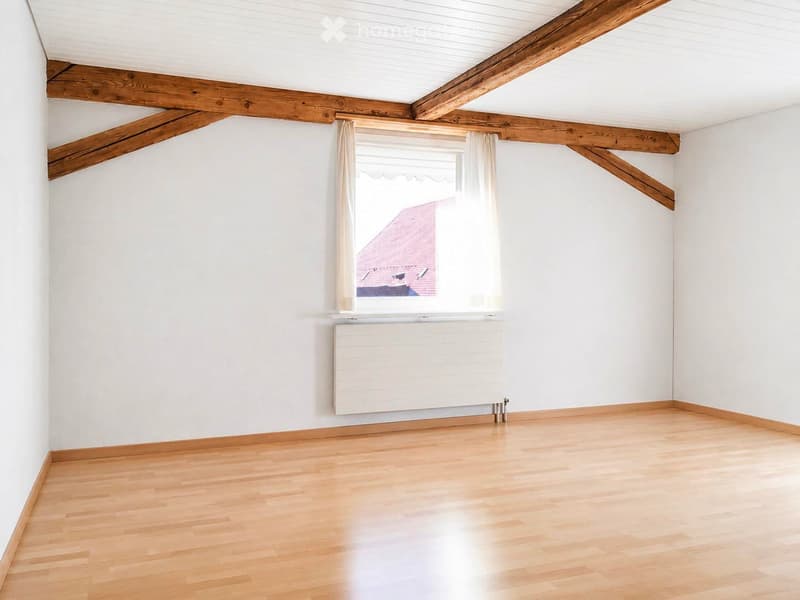 Empty room with wooden flooring, white walls, exposed wooden beams, a window with white curtains, and a radiator below the window.
