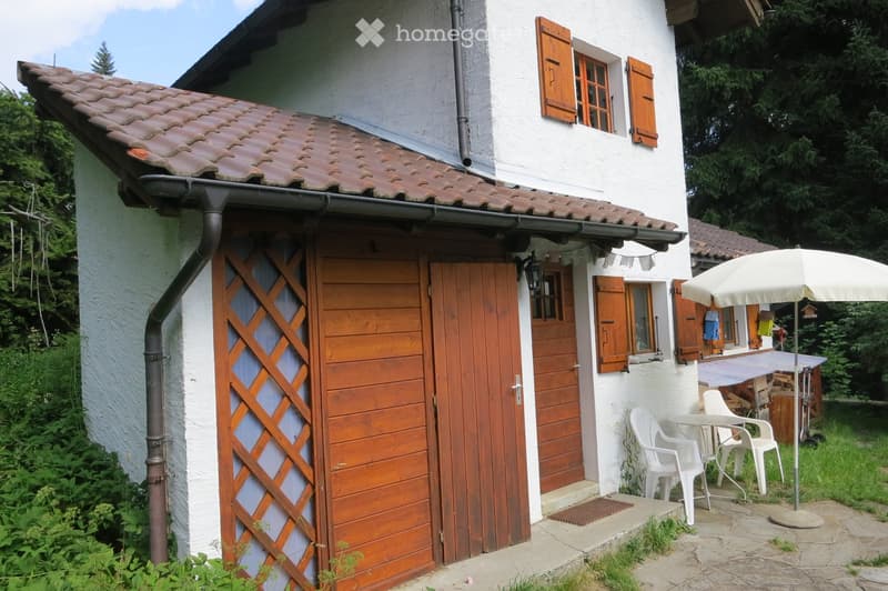 A white house with wooden shutters and a tiled roof, featuring a small patio area with white plastic chairs, a table, and an umbrella.