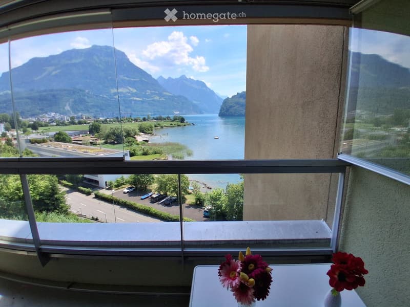 Balcony with large windows offering scenic views of a lake and mountains, decorated with a table and flower arrangements.