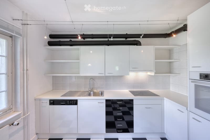 Modern kitchen with white cabinets, stainless steel sink, dishwasher, and oven. Features pendant lights and a window on the left side.