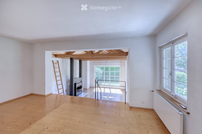 Empty room with wooden flooring, white walls, large windows, and a visible staircase leading to an upper level. No furniture or furnishings are present.