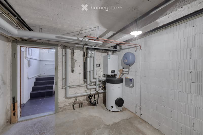 Utility room with exposed brick walls, concrete floor, and various pipes and water heaters. A staircase leads to another area.