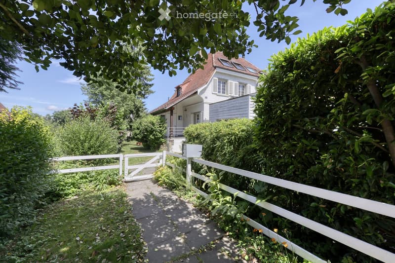 A white picket fence encloses a lush garden with various greenery and a pathway leading to a white house with a red-tiled roof and multiple windows.