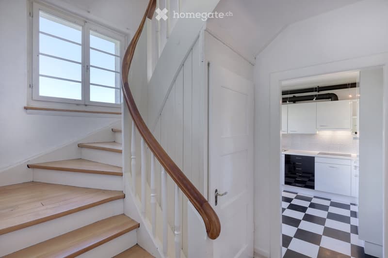 A narrow hallway with a wooden staircase leading upwards, featuring white walls and a large window on the left side. A door on the right opens into a modern kitchen with white cabinets, black countertops, and a checkered black-and-white floor.
