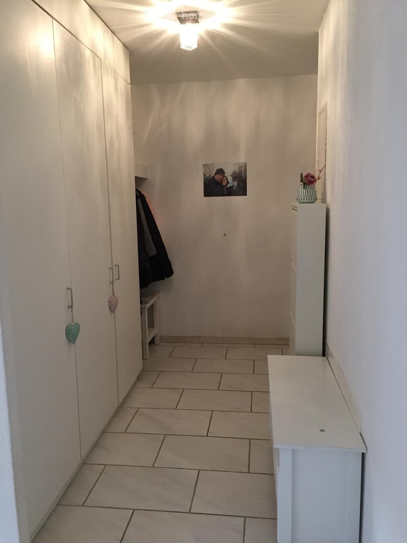 A narrow hallway with tiled flooring, white walls, and built-in storage cabinets. A small bench and a vase with flowers are visible on the right side.