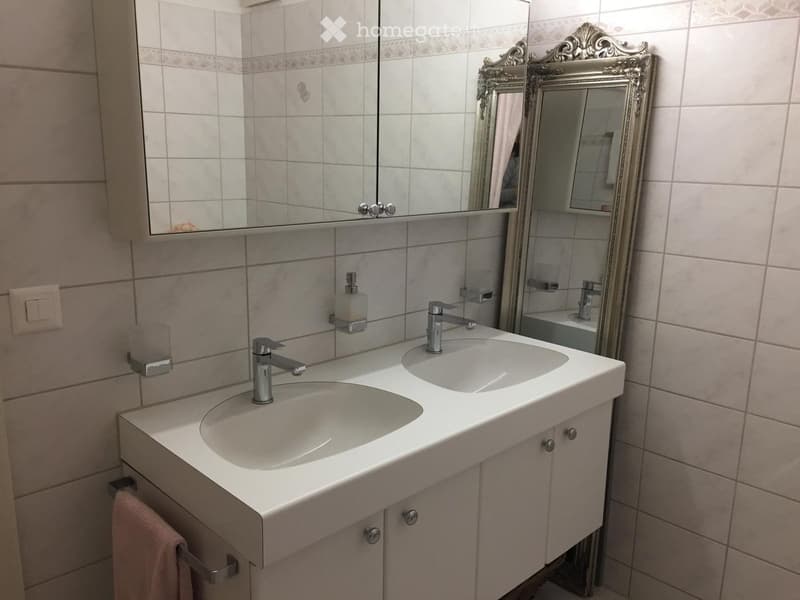 Bathroom with double sink, modern faucets, ornate mirror, and cabinetry.