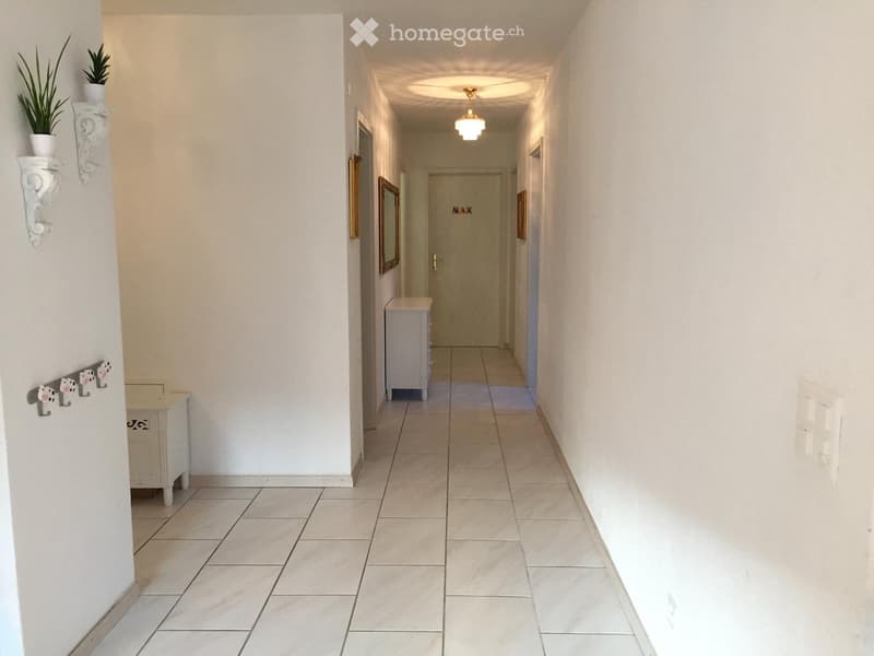 A clean, well-lit hallway with tiled flooring, white walls, and a ceiling light fixture. There are small decorative items on the walls and a closed door at the end.