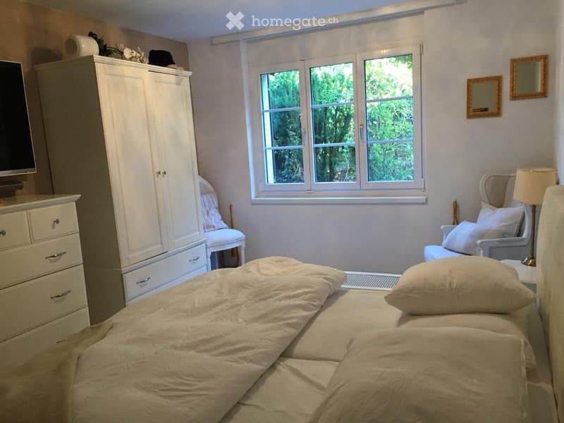 Cozy bedroom with a large bed, pillows, a wardrobe, a drawer unit, a chair, a table lamp, and a mirror on the wall.