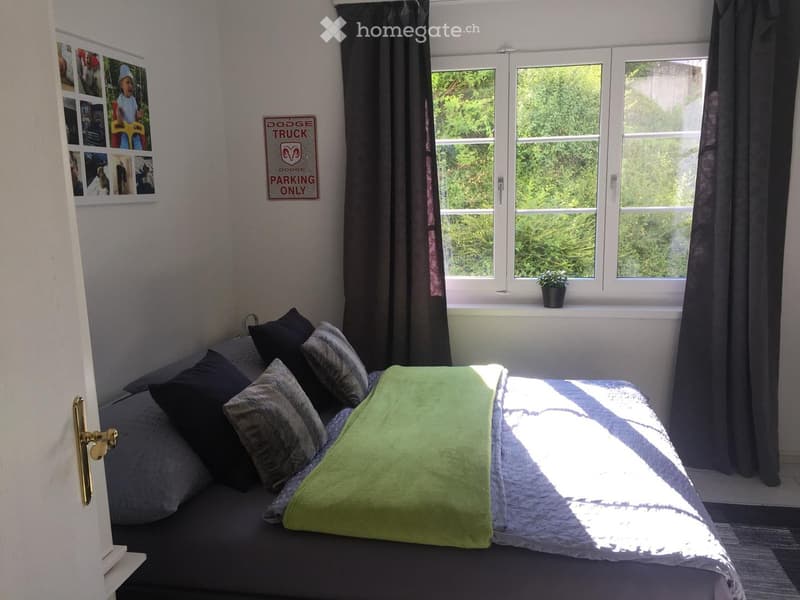 Cozy bedroom with a bed, pillows, a green blanket, and a window with a view of greenery. A small plant sits on the windowsill, and a sign on the wall reads "Dodge Truck Parking Only."