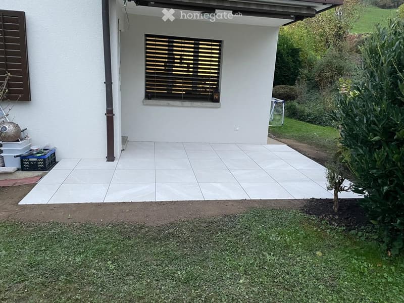 Outdoor patio area with tiled flooring, white walls, and a small garden with grass and plants. A window with blinds is visible on the house.