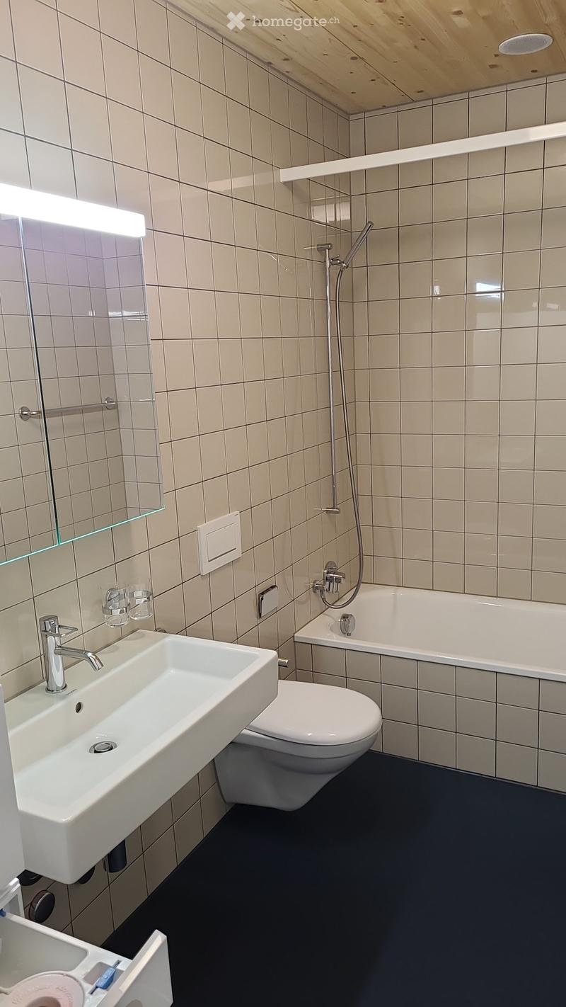 Modern bathroom with white tiles, a white sink, toilet, bathtub, and a glass shelf with a mirror above it. The floor is dark grey.