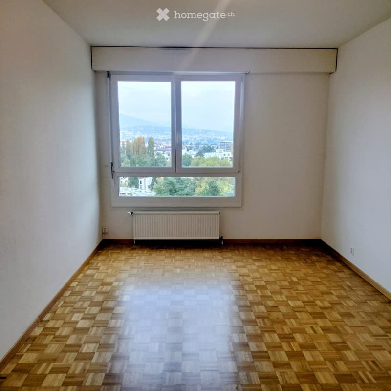 Empty room with wooden parquet flooring, white walls, large window offering a view, and a radiator below the window.