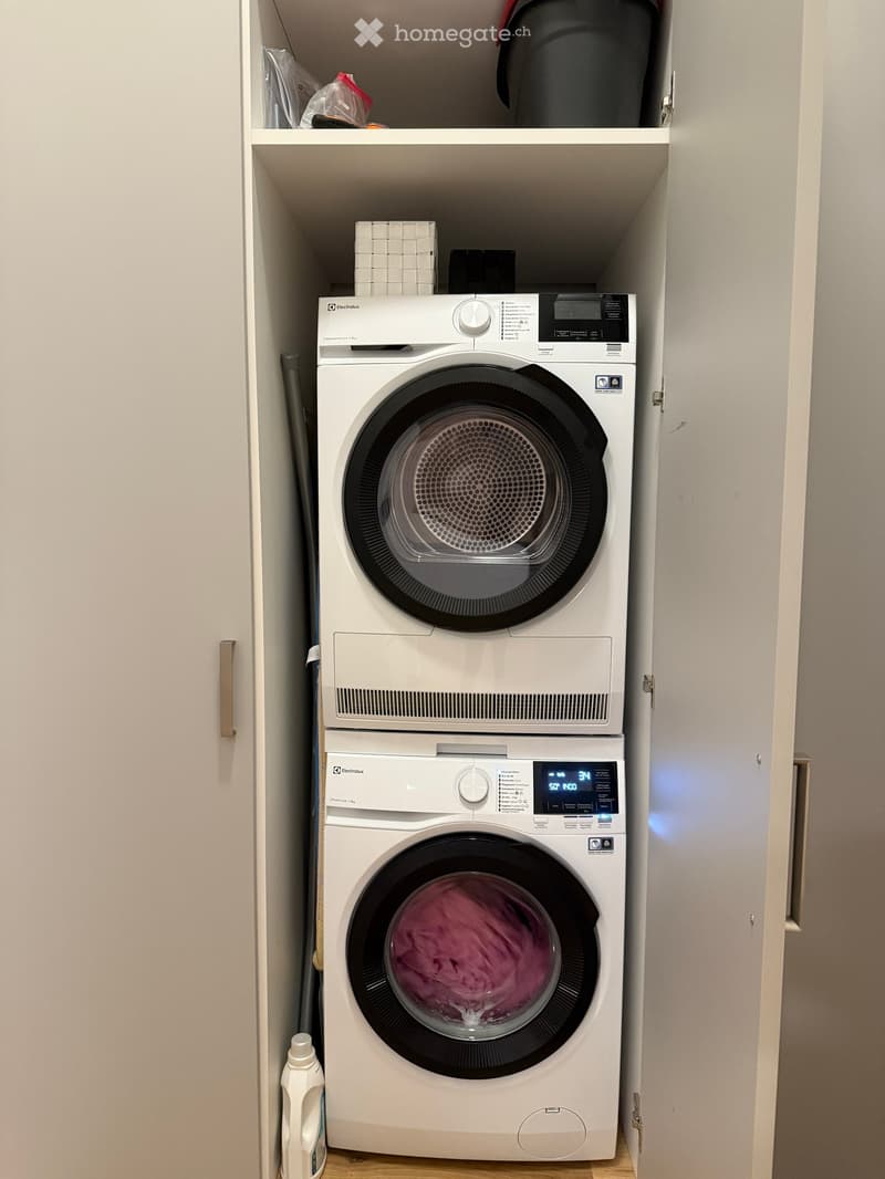 A compact laundry area with a white washing machine and dryer stacked vertically, a bottle on the floor, and shelves above.