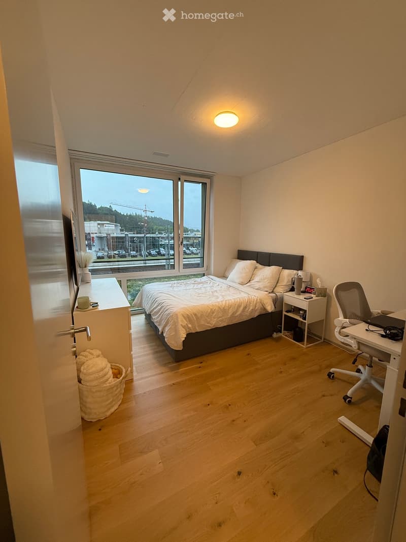 Modern bedroom with a large bed, nightstand, desk, and chair. Large windows offer a view of the outside area.