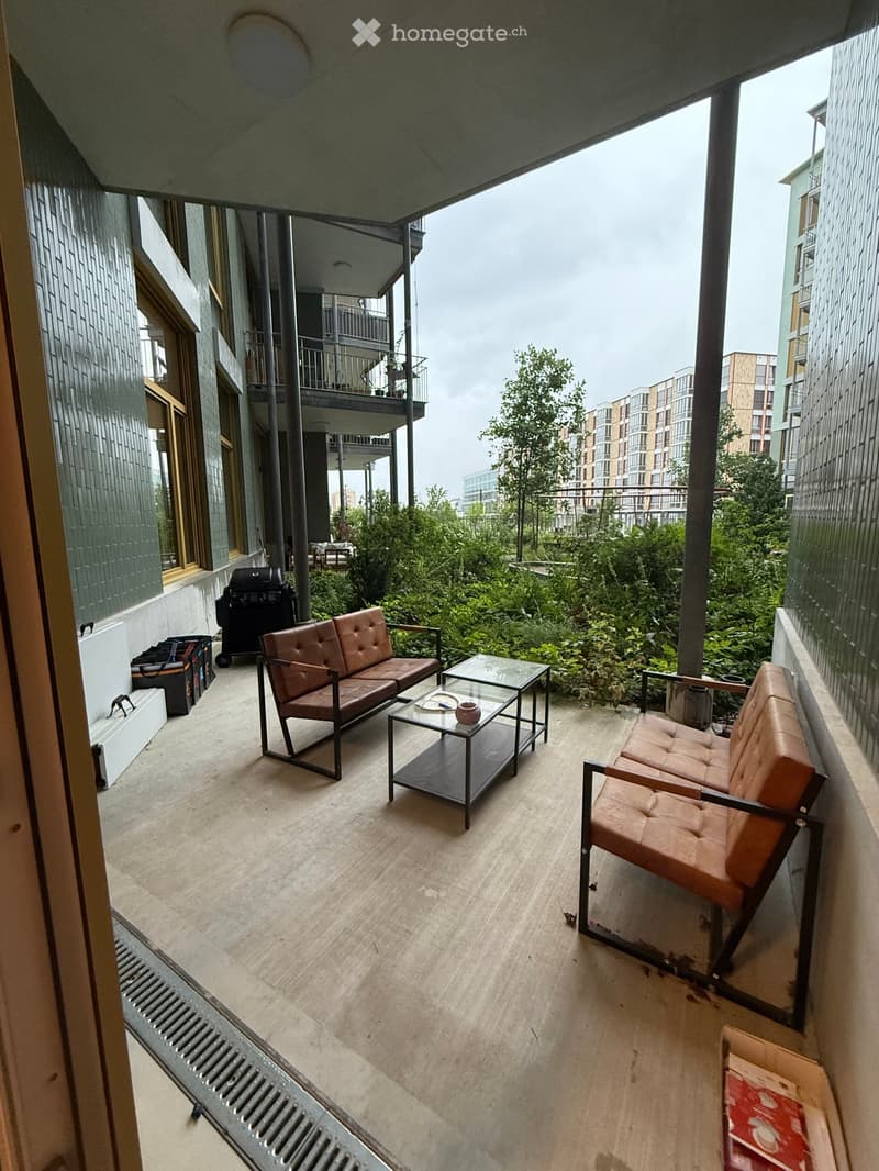 Modern balcony with concrete floor, two sofas, a glass coffee table, and a grill. Greenery and cityscape visible in the background.