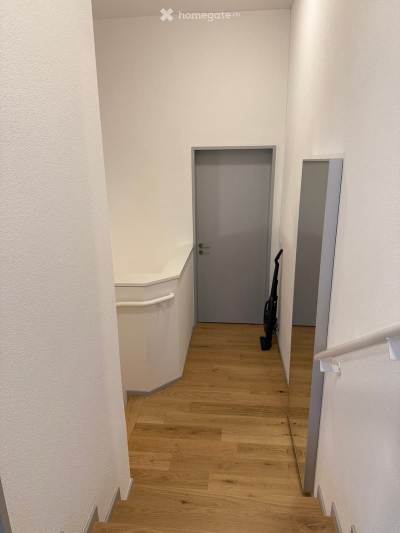 A narrow hallway with light wooden flooring, white walls, and a closed door at the end. A vacuum cleaner is placed against the wall near the door.