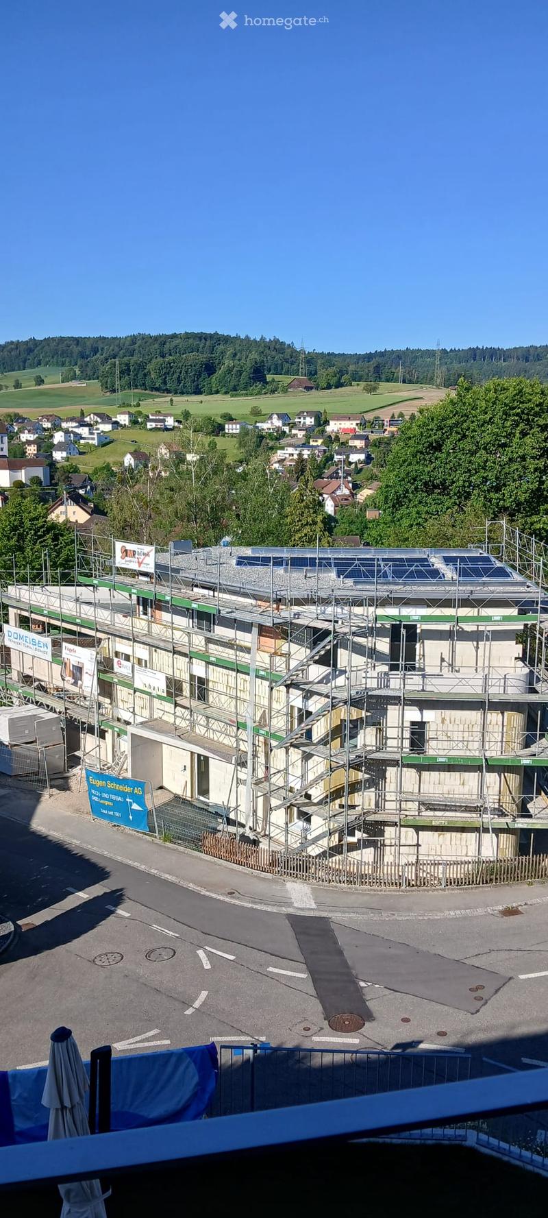 Multi-story building under construction with scaffolding, solar panels on the roof, and a fenced perimeter. The surrounding area includes a paved road, grassy fields, and scattered houses in the distance.