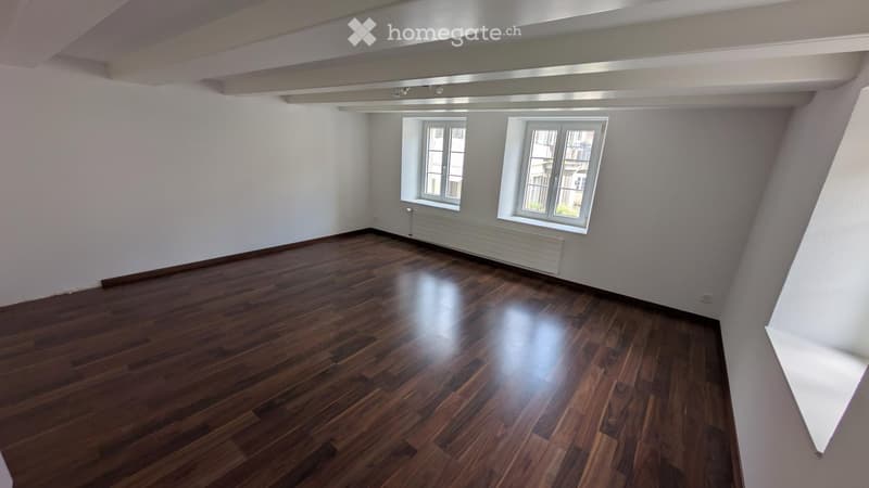 Empty room with wooden flooring, white walls, and two windows with white frames. The ceiling has exposed beams and lighting fixtures.