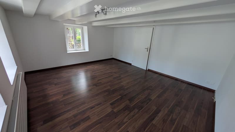 Empty room with wooden flooring, white walls, and a single window with a view of greenery outside. The room has a white door and exposed ceiling beams.