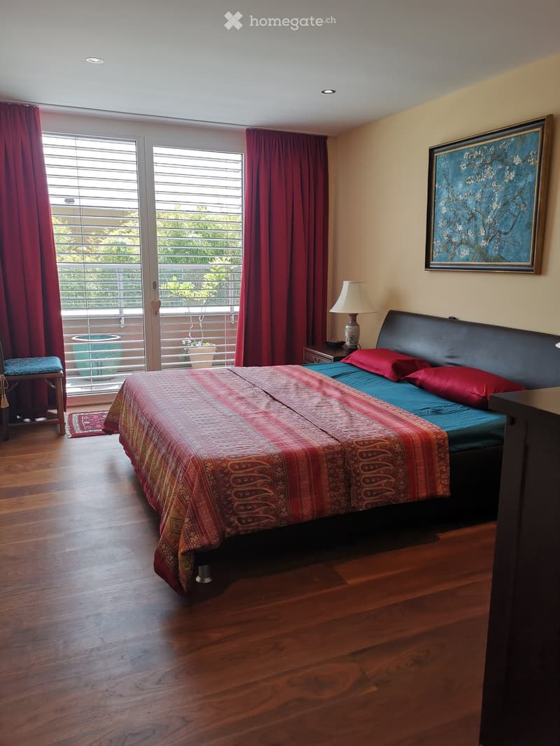 double bed, red and blue bedding, bedside tables, lamp, picture frame, wooden floor, sliding glass door, balcony, red curtains, rug, view of trees