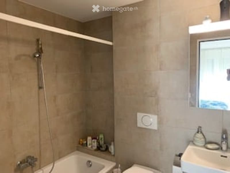 Modern bathroom with a bathtub, toilet, sink, mirror, and shower. The walls are tiled in a neutral color, and the space is well-lit with a built-in shelf for storage.