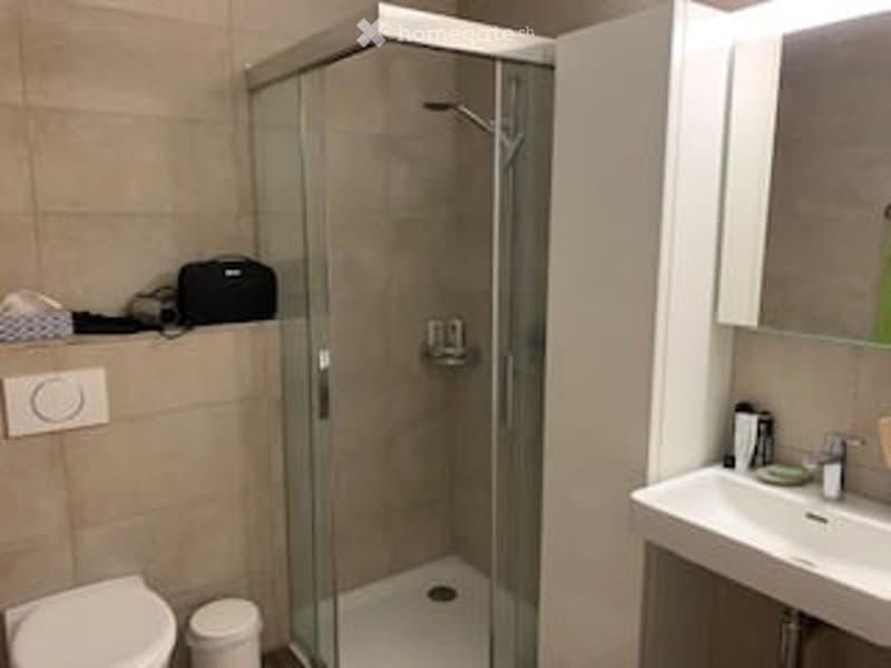 Modern bathroom with a glass-enclosed shower, toilet, sink, and mirror. The walls are tiled in a neutral color, and the floor is white.
