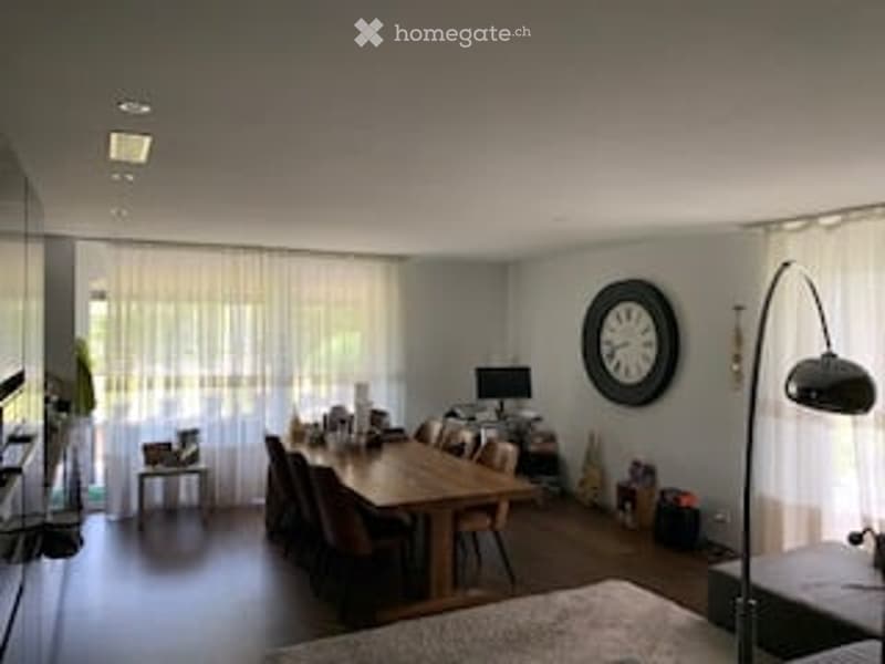 Bright, modern dining and living area with large windows, white curtains, a wooden dining table with chairs, a wall clock, a floor lamp, and a small side table.