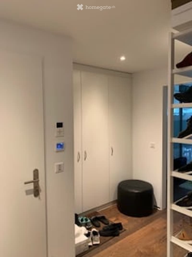 A small, modern room with a white wardrobe, a black round stool, and a wooden floor. There are some shoes and items on the floor near the door.