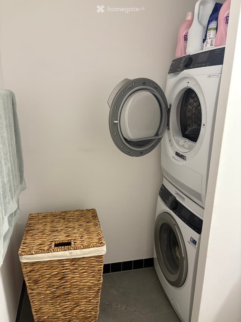 Compact bathroom with a stacked washing machine and dryer unit, a wicker basket, and a towel hanging on the wall.