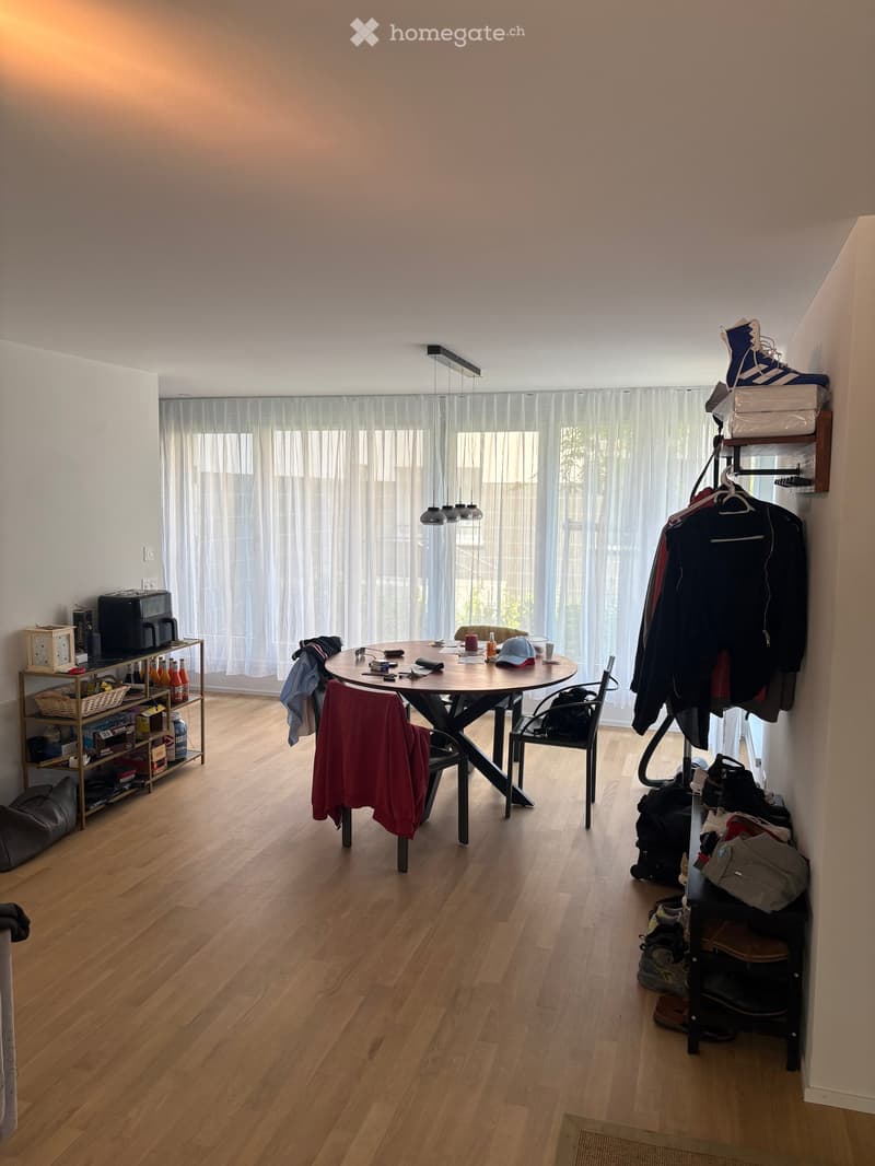 A modern dining area with a round wooden table, chairs, a shelf with various items, and a hanging lamp. The room features wooden flooring and large windows with white curtains.