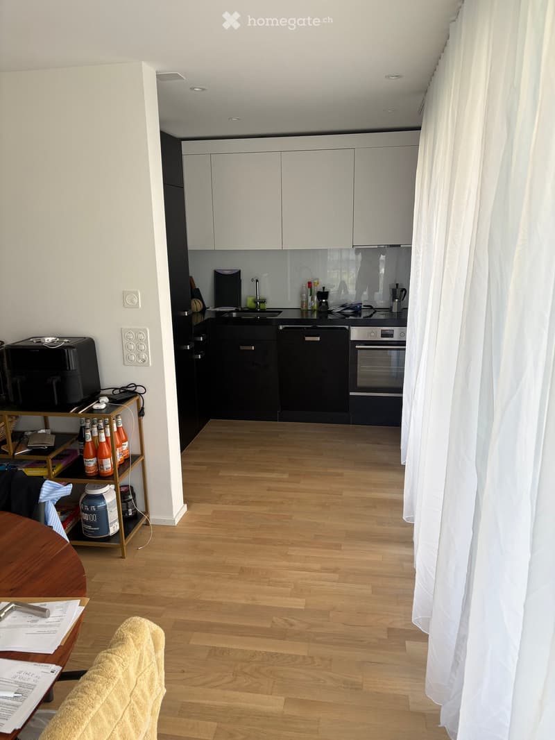 A modern, open-plan living and kitchen area with light wooden flooring, white cabinets, and a black oven. A small shelf holds several bottles and a container, and a sofa is partially visible in the foreground.