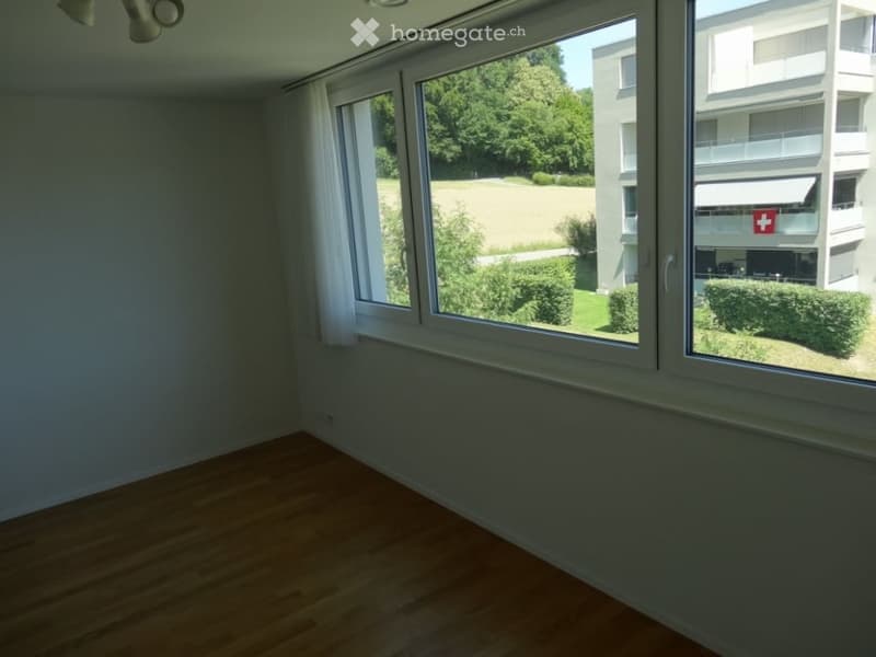 Empty room with wooden flooring and large windows offering a view of greenery and a building with a Swiss flag.