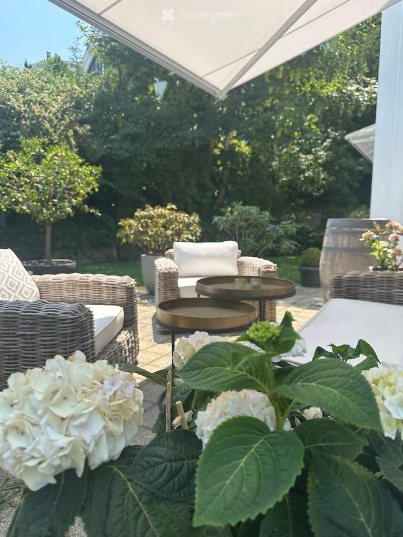 A well-maintained garden with wicker chairs, a small table, a barrel, and lush greenery, including flowering plants and trees.