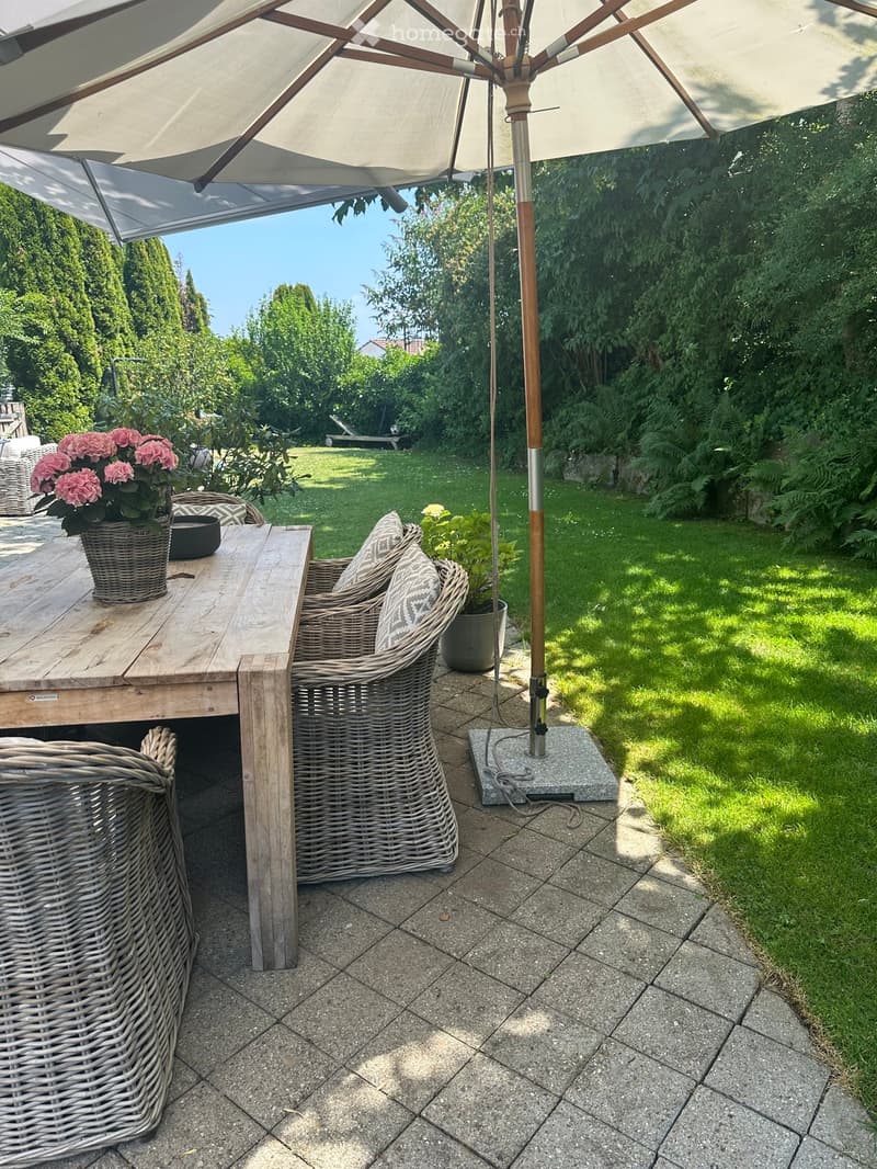 Garden patio with a wooden table, wicker chairs, umbrella, and flower pot.