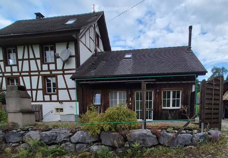 Two-story house with a traditional half-timbered design and a separate wooden outbuilding, surrounded by a stone wall and greenery.
