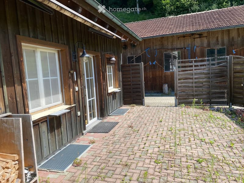 Brick-paved outdoor area with wooden structures, a gate, and a stack of firewood near the building entrance.