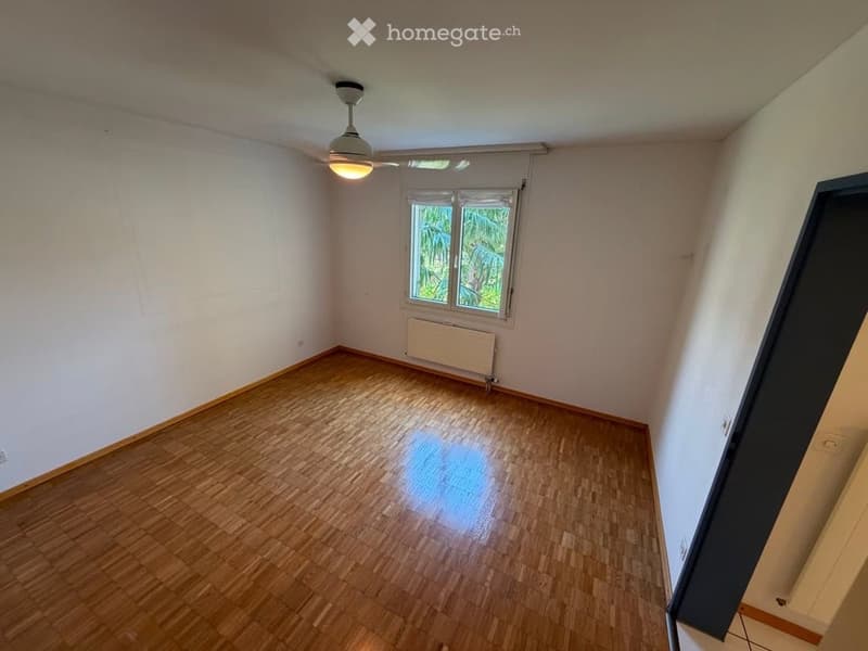 Empty room with wooden flooring, white walls, a ceiling fan with light, and a window with a view of greenery outside.