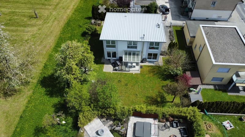 Aerial view of a modern two-story house with a flat roof, surrounded by a well-maintained lawn and trees. The house features large windows and a covered patio area with outdoor furniture. Adjacent to the house is a smaller building with a flat roof and so